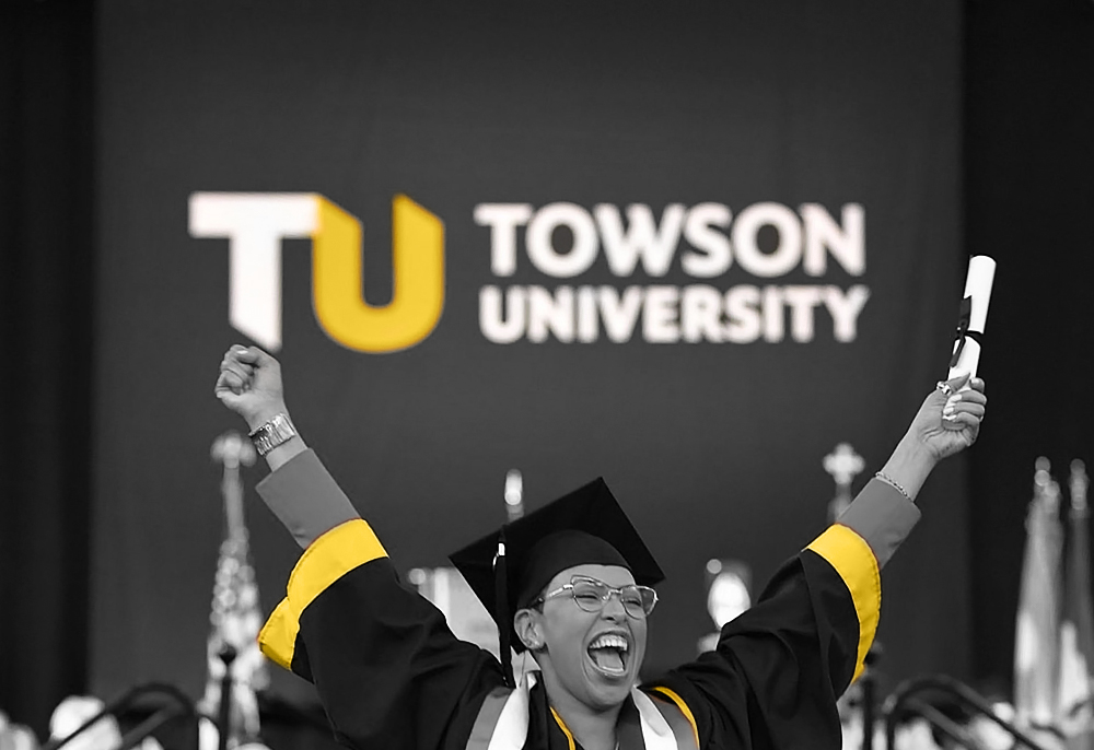 women in graduation gown and cap holing up her degree and cheering in front of a TU - Towson University banner